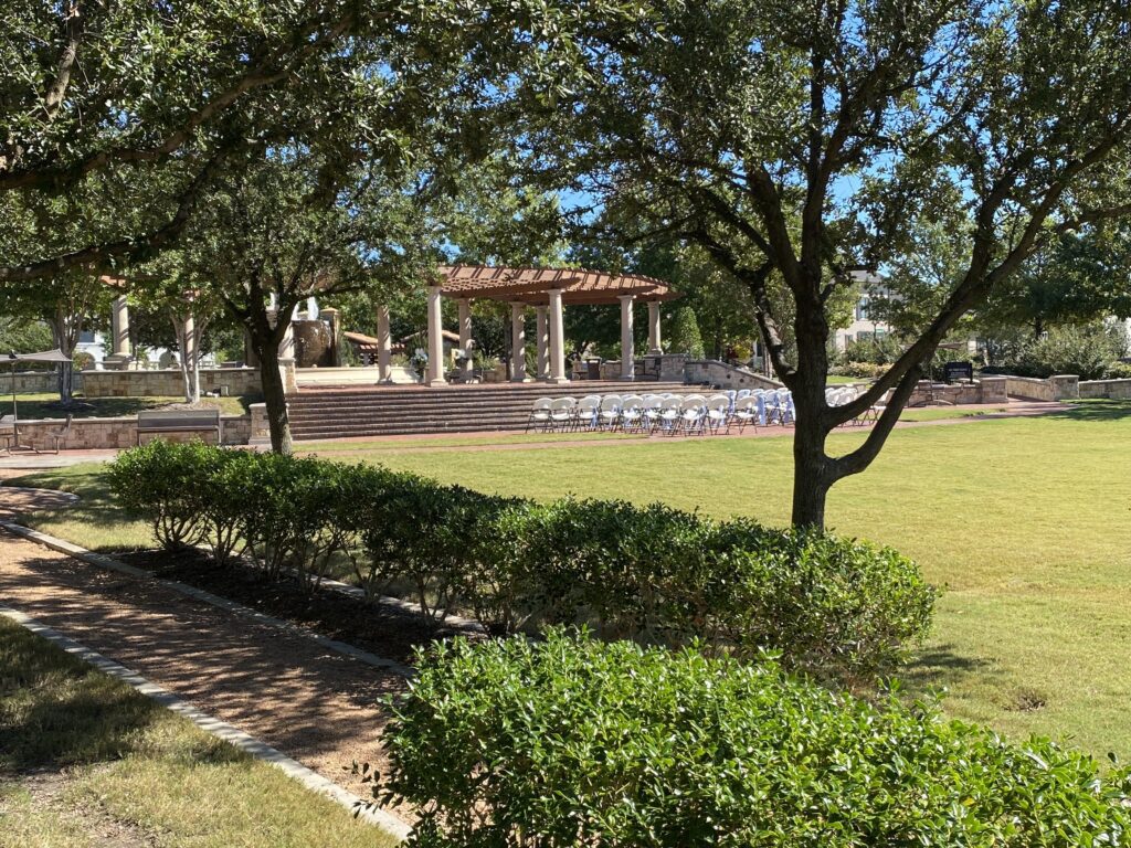 Outdoor pavilion and seating area in a sunny park surrounded by trees and greenery.